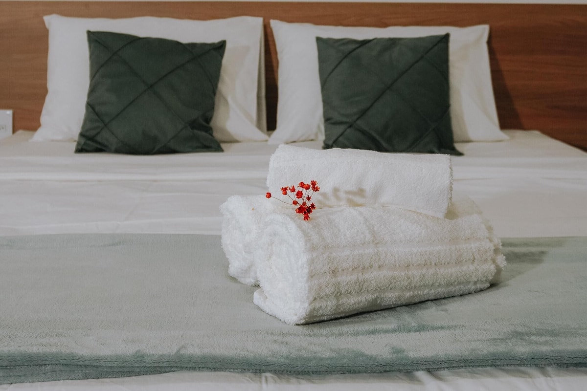 A neatly made bed with white linens, two green pillows, and a white towel with red flowers.
