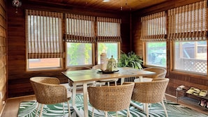 Un interior de madera con una mesa y sillas, ventanas amplias con persianas, un ventilador de techo y una planta.