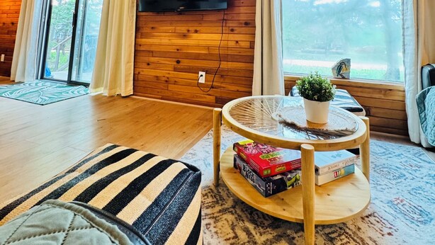 Interior acogedor de una cabaña de madera con un televisor grande montado en la pared, una mesa redonda de madera con una planta y libros, y un cojín a rayas en el suelo.