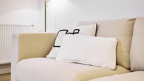 Een beige bank met twee kussens, een effen en een met een zwart dessin, in een kamer met een vloerlamp en een radiator.