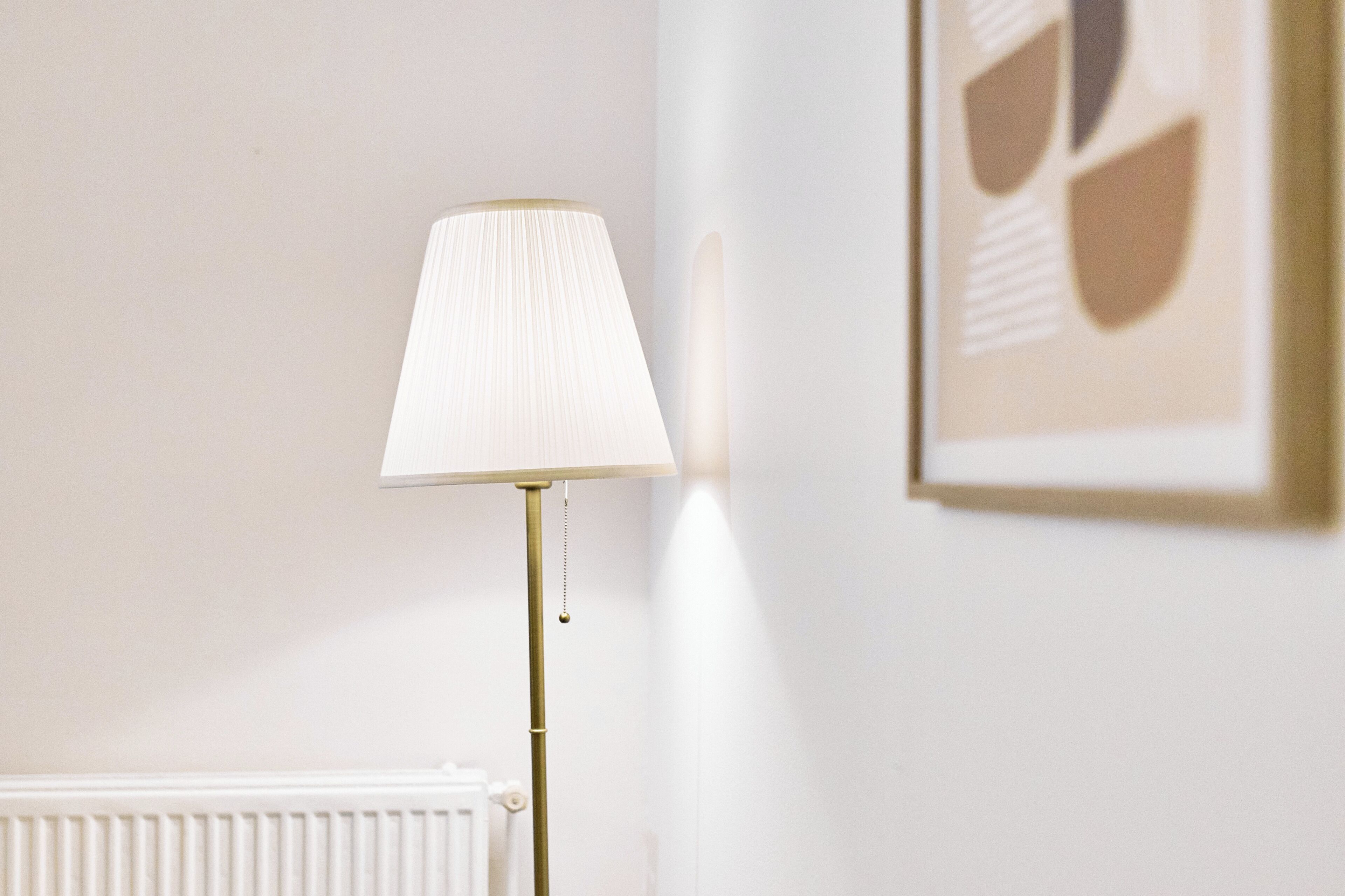 Een vloerlamp met een witte lampenkap in een kamer met een witte radiator en een lijst met abstracte kunst aan de muur.