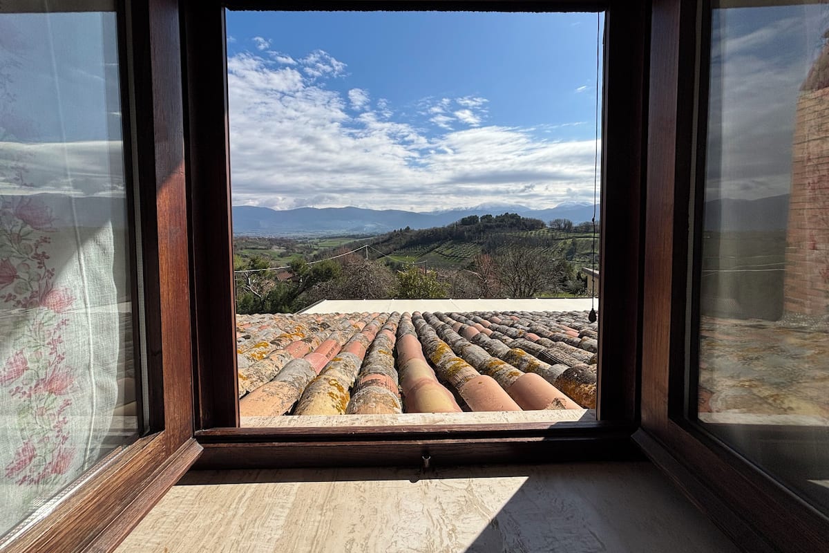 Une vue depuis une fenêtre donnant sur un toit recouvert de tuiles, avec un paysage vallonné et un ciel partiellement nuageux en arrière-plan.