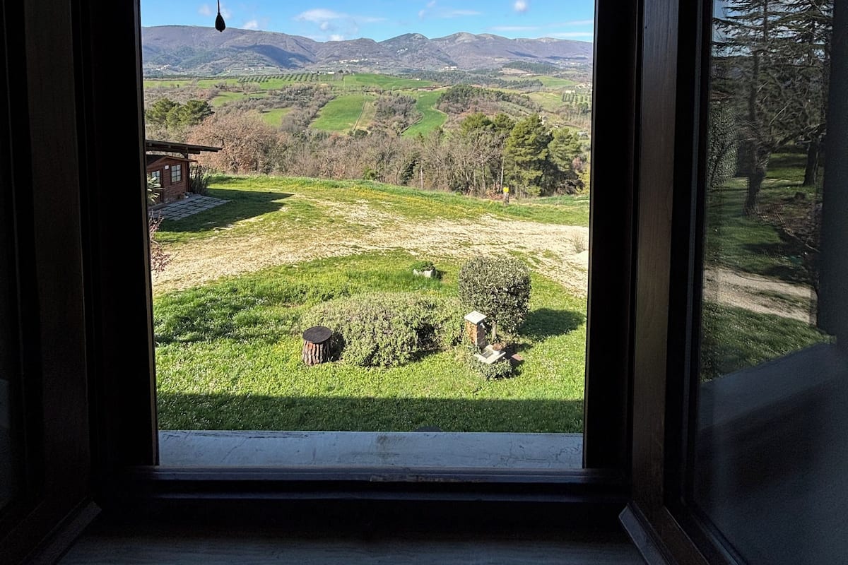 Une pièce dotée d’une grande fenêtre donnant sur un paysage verdoyant, avec des collines et des arbres.