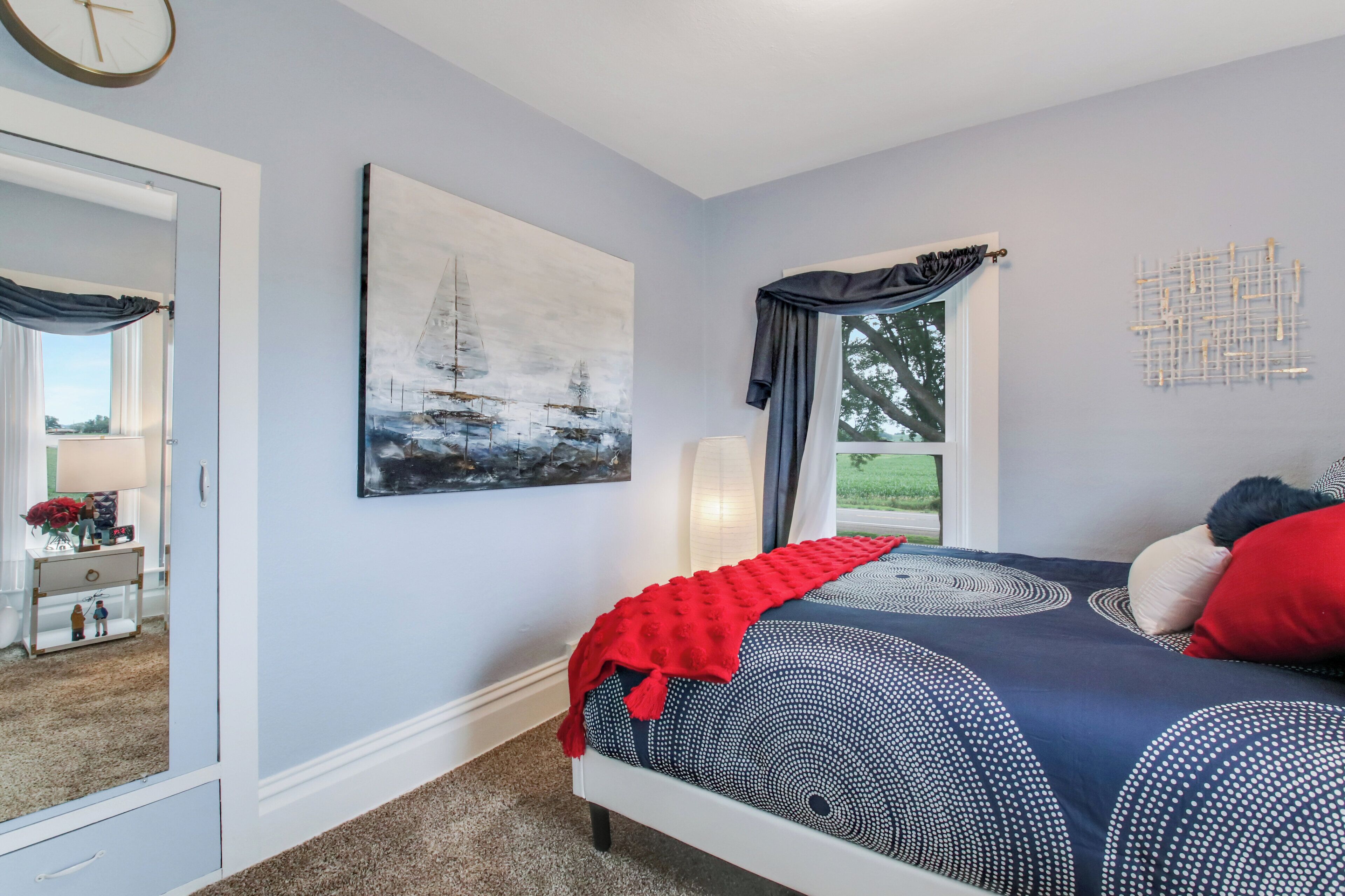 A bedroom with a bed, a painting, a window with curtains, and a wall-mounted clock.