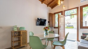 A dining area with a round table, green chairs, a coffee machine, a TV on the wall, and a view through an open door. - La Casa dei Fiori d'Arancio (Capoterra)