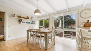 Ruime eethoek met een houten tafel en witte stoelen, grote ramen met uitzicht op de omgeving, en een plafond met zichtbare balken.