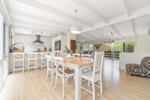 A spacious dining area with a wooden table and white chairs, a kitchen with modern appliances, and a large window offering a view of the outdoors.