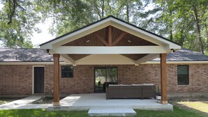 A covered patio area with a built-in bench, a brick house in the background, and a grassy lawn. - Welcome to your perfect getaway or family retreat in beautiful Magnolia Texas ! (Magnolia)
