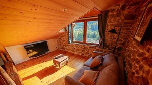 A cozy cabin interior with a stone wall, wooden ceiling, a TV, a sofa, and a window overlooking a forest. - Characteristic village house, sleeps 10 (Beauvezer)