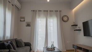 A modern living room with a grey sofa, a small table with a tray, a TV on the wall, and sheer curtains. - Seaside Charm in Palaio Faliro – Walk to Beach & Cafés! (Paleo Faliro)