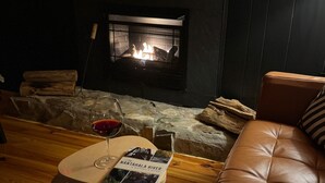 A cozy living room with a fireplace, a leather sofa, and a small table with a glass of red wine and a magazine. - Angler's Chalet (Topton)