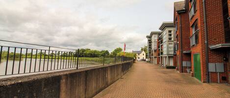A brick pathway beside a river with a metal railing, leading to a row of brick buildings with green doors.