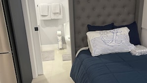 Une chambre à coucher avec une tête de lit capitonnée grise, un lit avec une literie bleu marine et un tapis blanc menant à la salle de bain.