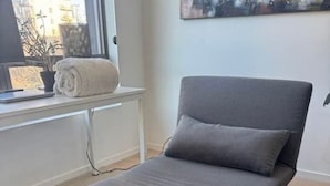Un lit simple avec un matelas gris et des oreillers assortis, placé contre un mur blanc, avec une fenêtre sur le côté gauche.