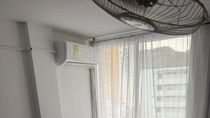 A bedroom with a single bed, a ceiling fan, an air conditioning unit, and a window with sheer curtains.