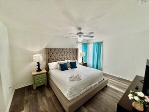 A bedroom with a tufted headboard, a bed with pillows, a nightstand with lamps, a ceiling fan, and a TV on a stand.