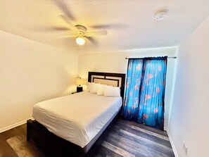 Habitación de hotel con cama, ventilador de techo, mesita de noche con lámpara y ventana con cortinas.
