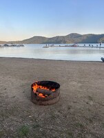 Una playa con un brasero en primer plano y barcos en el agua al fondo.