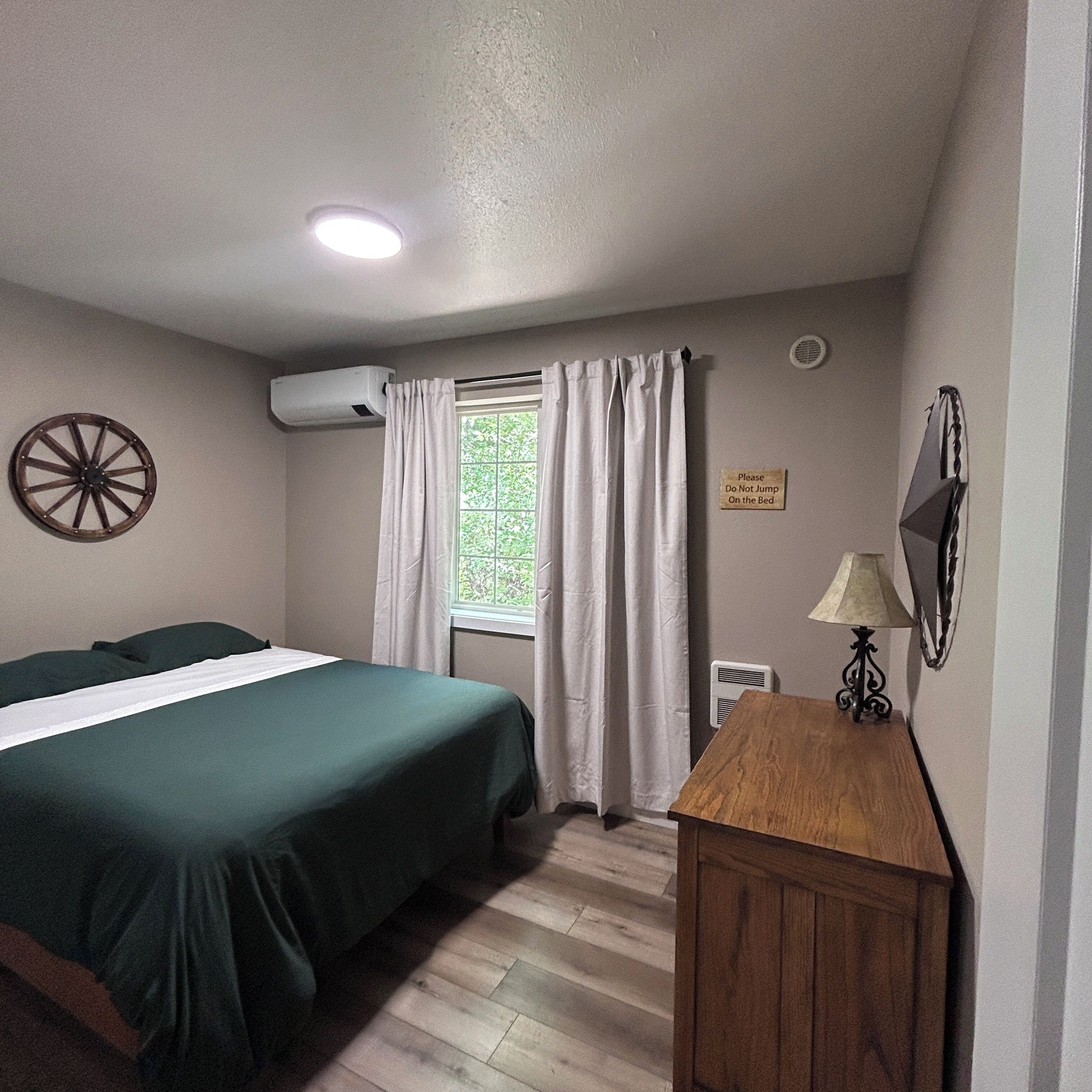Une chambre avec un lit, une table de chevet en bois, une lampe fixée au mur, une fenêtre avec des rideaux, un appareil de climatisation et un panneau sur le mur.