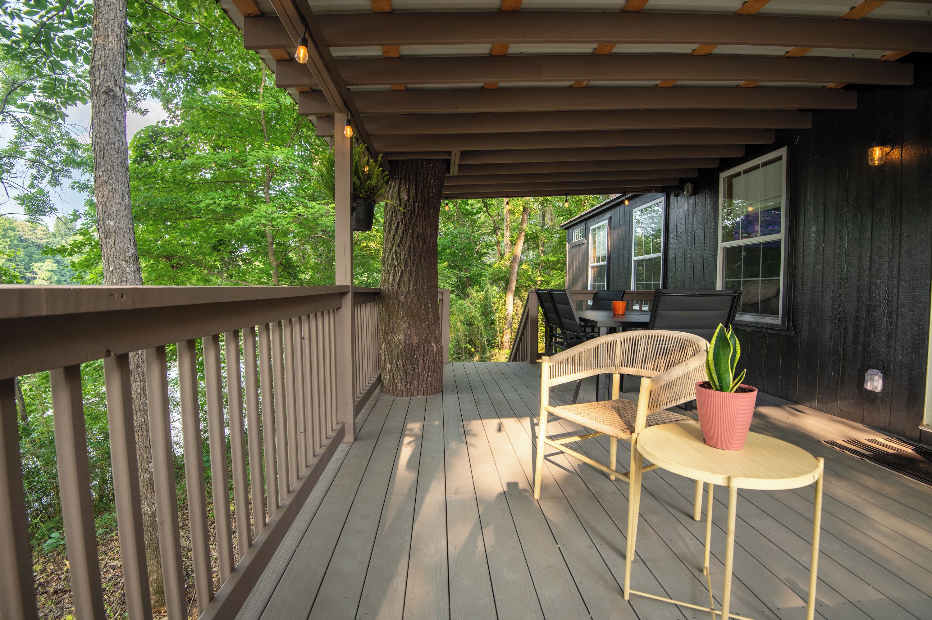 Eine Holzterrasse mit Tisch und Stühlen, einem Blumentopf und Blick auf ein Haus durch ein Fenster.