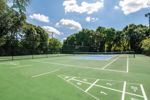 Un court de tennis en plein air, doté d’un filet, entouré d’arbres et surplombé d’un ciel dégagé.