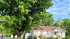 A white single-story house with a red roof, a porch, and a small front yard with a tree and a picnic table.