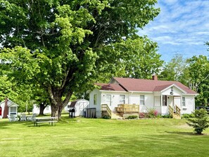 Et hvidt etplanshus med rødt tag, en veranda og en lille forhave med et træ og et picnicbord.