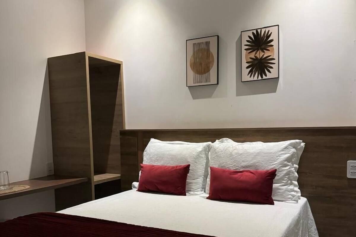 Ein ordentlich bezogenes Bett mit weißen Laken und roten Kissen, ein Holzkopfstück und zwei Wandbilder.