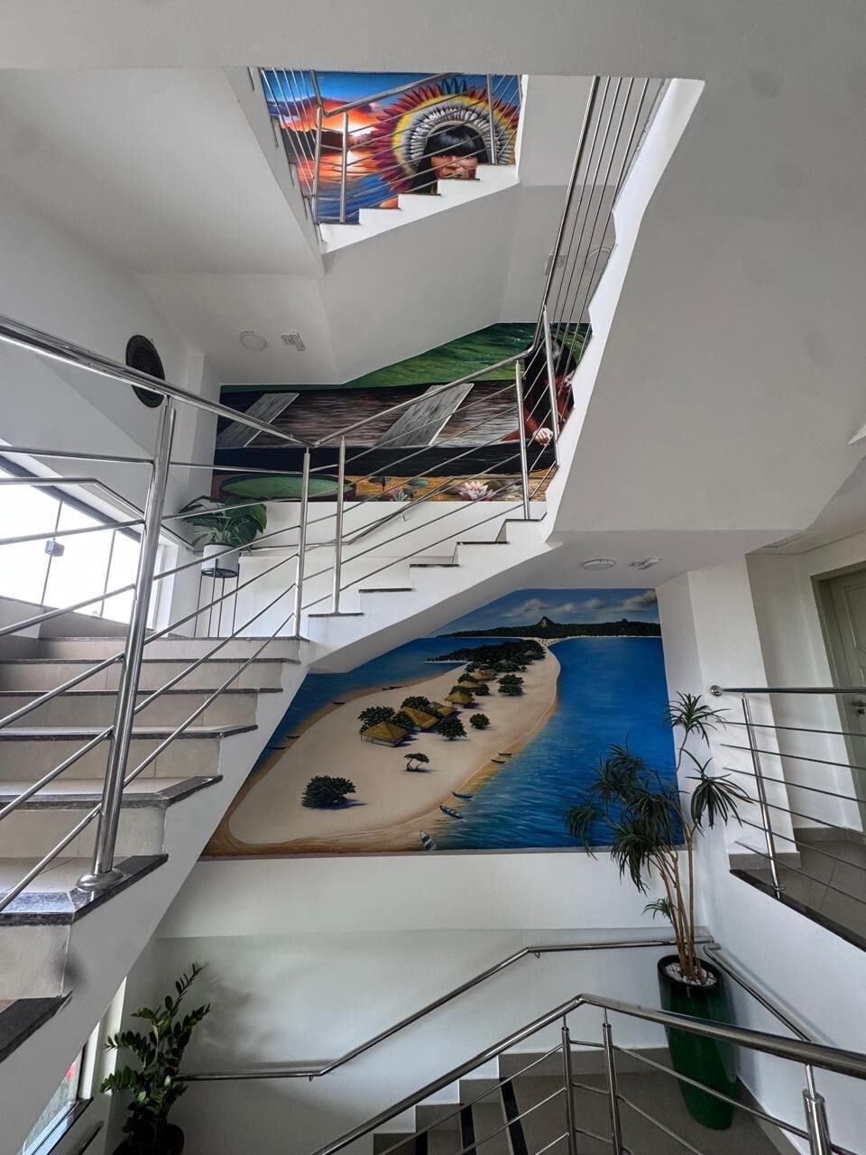 Interior com vários níveis, incluindo uma escadaria, um mural com uma cena de praia e plantas em vasos.