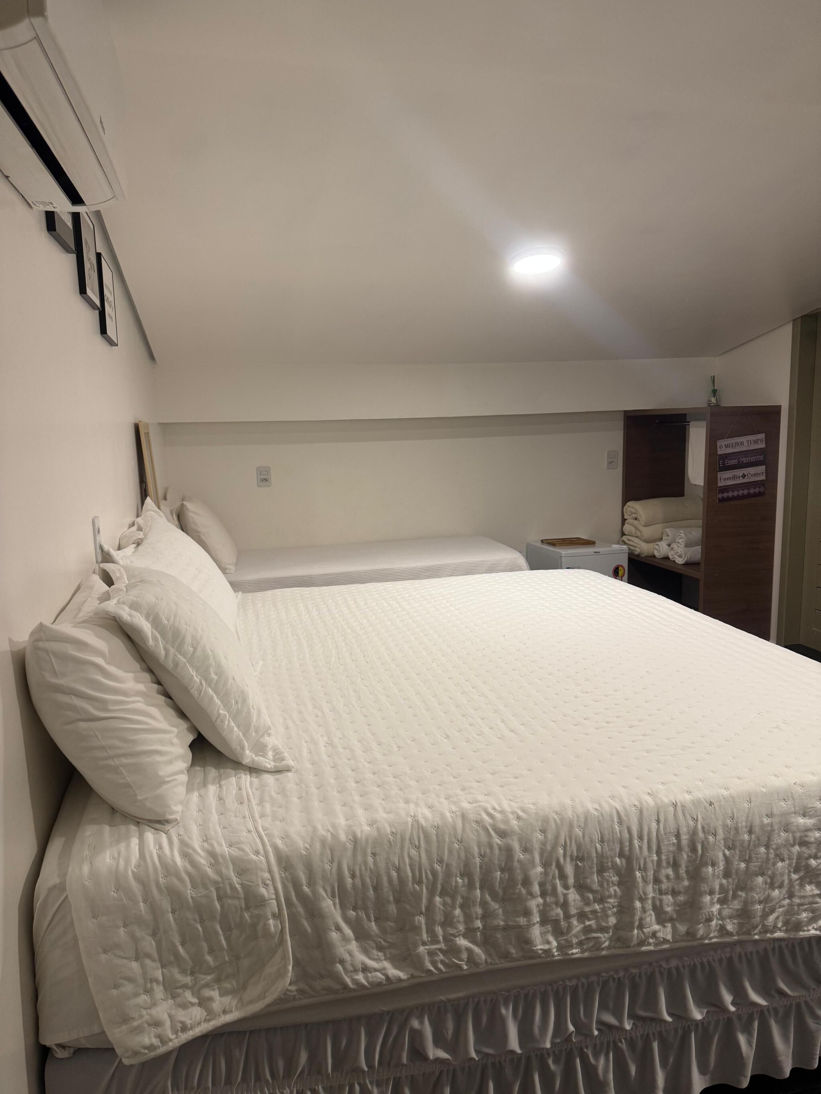 A single-bed room with a white bedspread, pillows, a bedside table, a nightstand, and a wall-mounted air conditioner.
