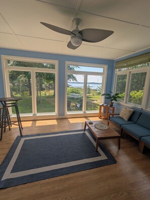 A room with a ceiling fan, large windows with a view of a lake, a blue sofa, and a dining table with chairs.