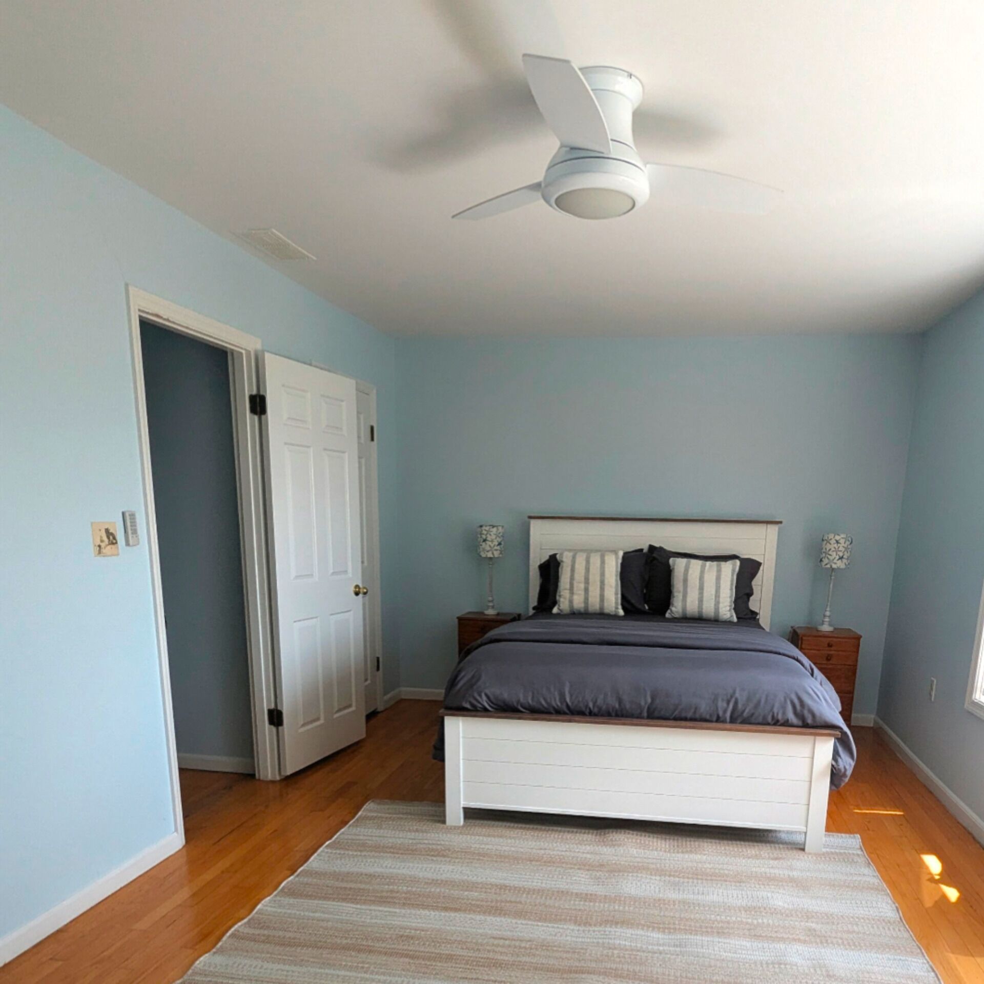 A bedroom with a bed, a ceiling fan, a door, and a nightstand with a lamp.