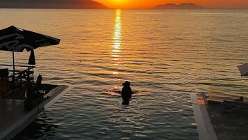 A sunset over a calm body of water with a person standing in the water, a dock with umbrellas, and a distant mountain range.