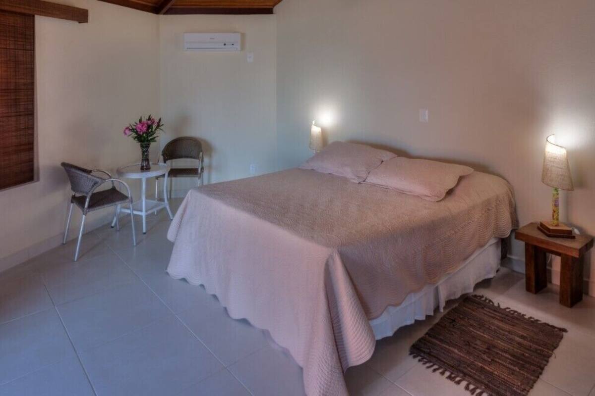 Ein Schlafzimmer mit einem Bett, zwei Stühlen, einem kleinen Tisch mit Blumenvase, einer Lampe und einem Fenster mit Jalousien.