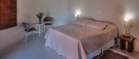 A bedroom with a bed, two chairs, a small table with a vase of flowers, a lamp, and a window with blinds.