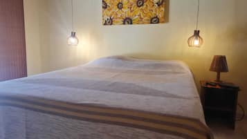 Cama bem arrumada com manta listrada, duas luminárias pendentes, pintura de flores na parede e uma pequena mesa de cabeceira com abajur.