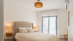 Ein Schlafzimmer mit einem gesteppten Kopfteil, einem großen Bett mit beiger Bettdecke, zwei Nachttischlampen, einer Korblampe und Blick ins Freie durch ein Fenster mit transparenten Vorhängen.