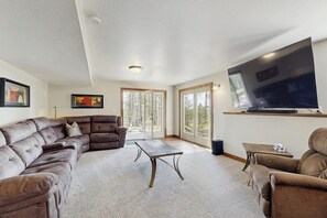 A spacious living room with a large flat-screen TV, leather sofas, a coffee table, and large windows offering a view of trees. - Spacious and secluded home with mountain views, gas grill, decks & home office (Rapid City)