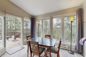 A room with a large table and chairs, a floor lamp, and a view of the outdoors through large windows.