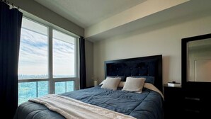 A bedroom with a large window, a dark headboard, and a bed with a blue bedspread and two pillows. - Modern Designed Home (Sebright)