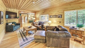 Een gezellig houten chaletinterieur met houten wanden en vloeren, een woonkamer met leren banken en een flatscreen televisie, en een groot raam met uitzicht op een bos.