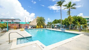 Une piscine dotée d’une structure de toit en chaume, de palmiers et, au fond, d’un bâtiment portant l’inscription « BBQ & Pizza ».
