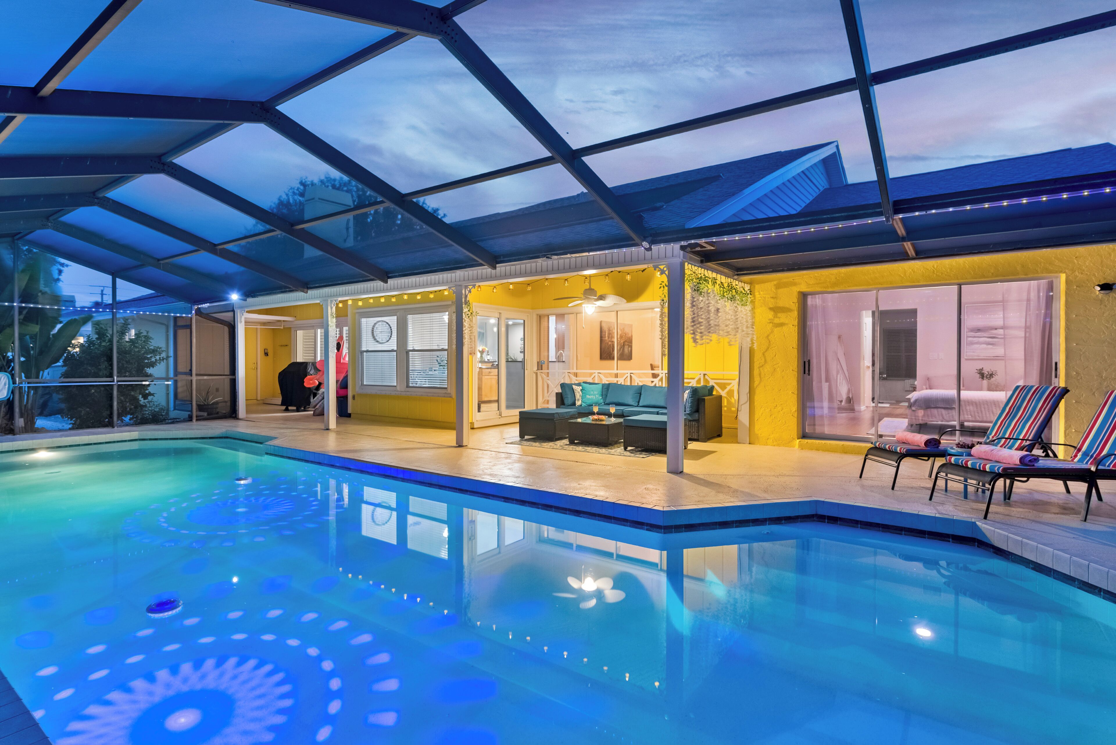 Une piscine dotée d’un toit en verre, un solarium aux murs jaunes et agrémenté d’un canapé, et un espace patio avec des chaises longues.