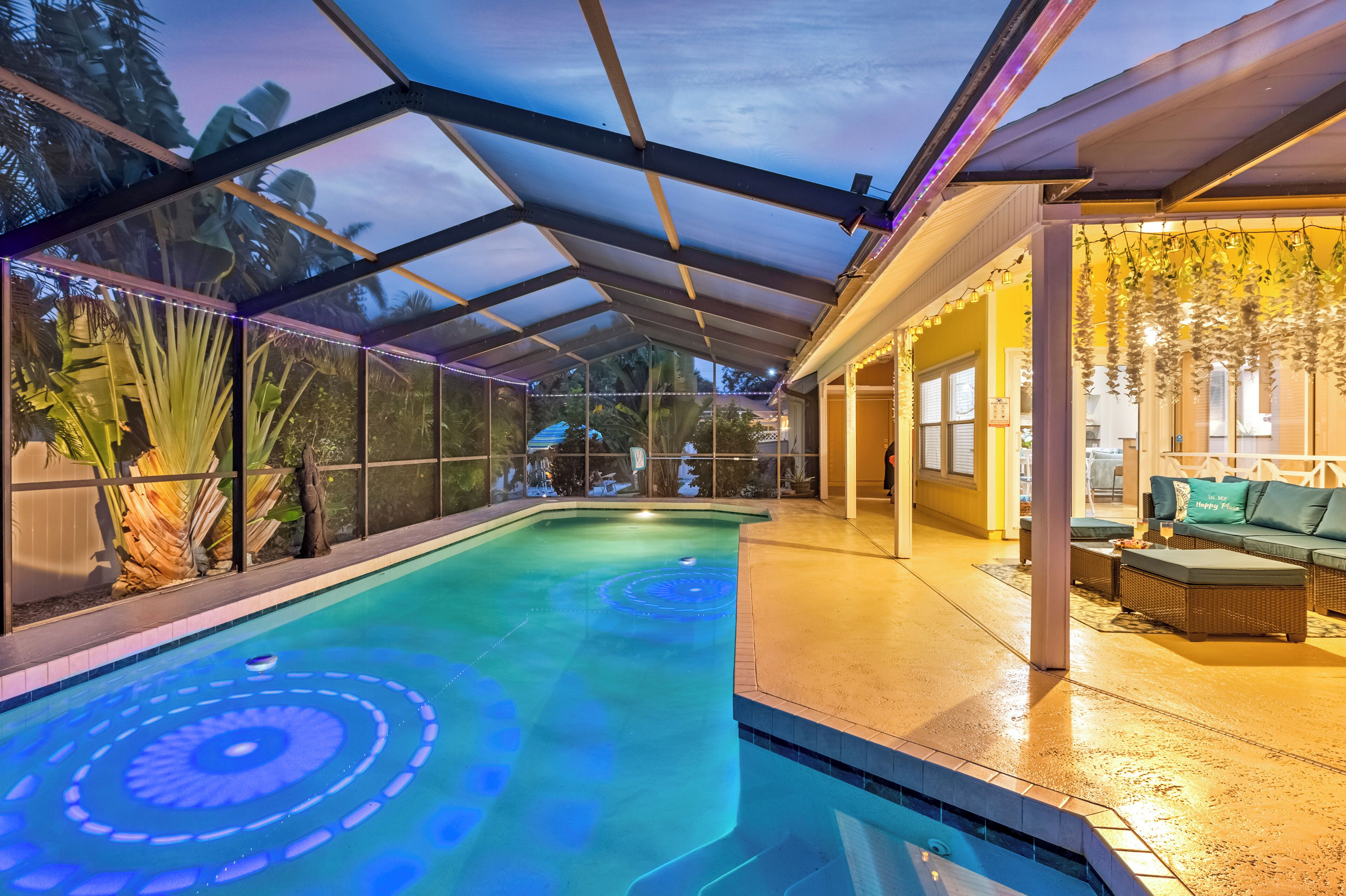An indoor swimming pool with a glass ceiling, surrounded by plants and a seating area with cushions.