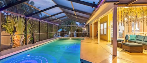 An indoor swimming pool with a glass ceiling, surrounded by plants and a seating area with cushions.