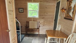A cozy room with a wooden floor, a dining table with chairs, a window with a view of greenery, and a door leading to another area.
