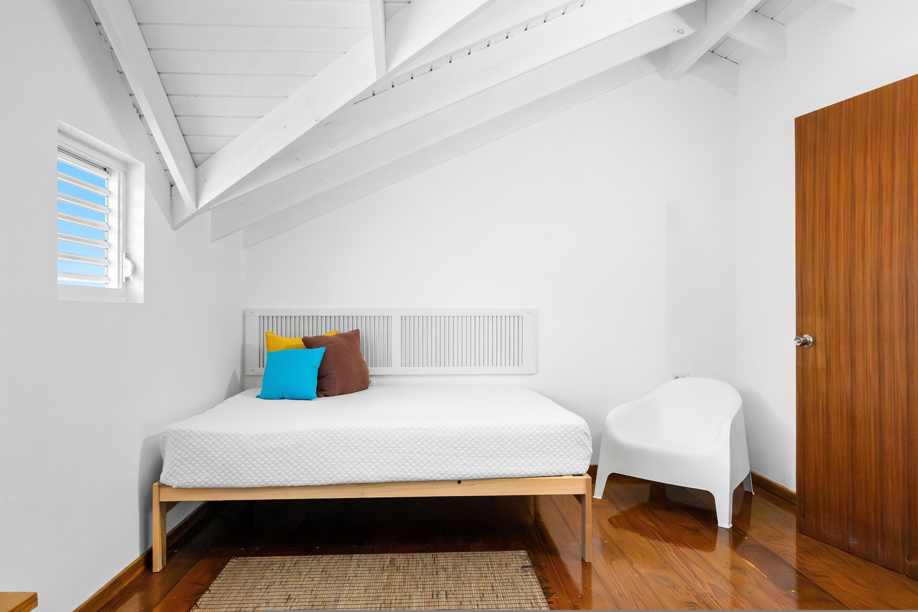 Un dormitorio con una cama, una silla blanca, una puerta de madera y una ventana con persianas.