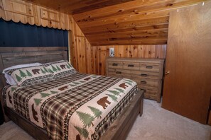 A bedroom with a wooden ceiling, a bed with a forest-themed bedspread, a dresser, and a door. - Cozy 3-bedroom log Cabin.  Just minutes away from Bass Lake. (Oakhurst)
