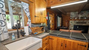 A wooden cabin kitchen with a large sink, wooden cabinets, a stone fireplace, and a window with a view of greenery.