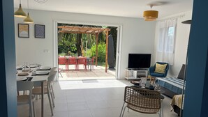 Een moderne woonkamer met een televisie, bank, eethoek en uitzicht door een glazen deur op een buitenruimte.